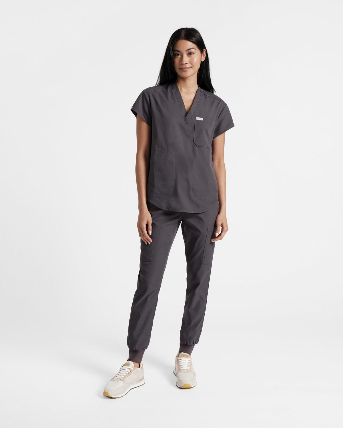 Woman in dark grey scrubs with a v-neck shirt that has a rounded hem and side pockets.