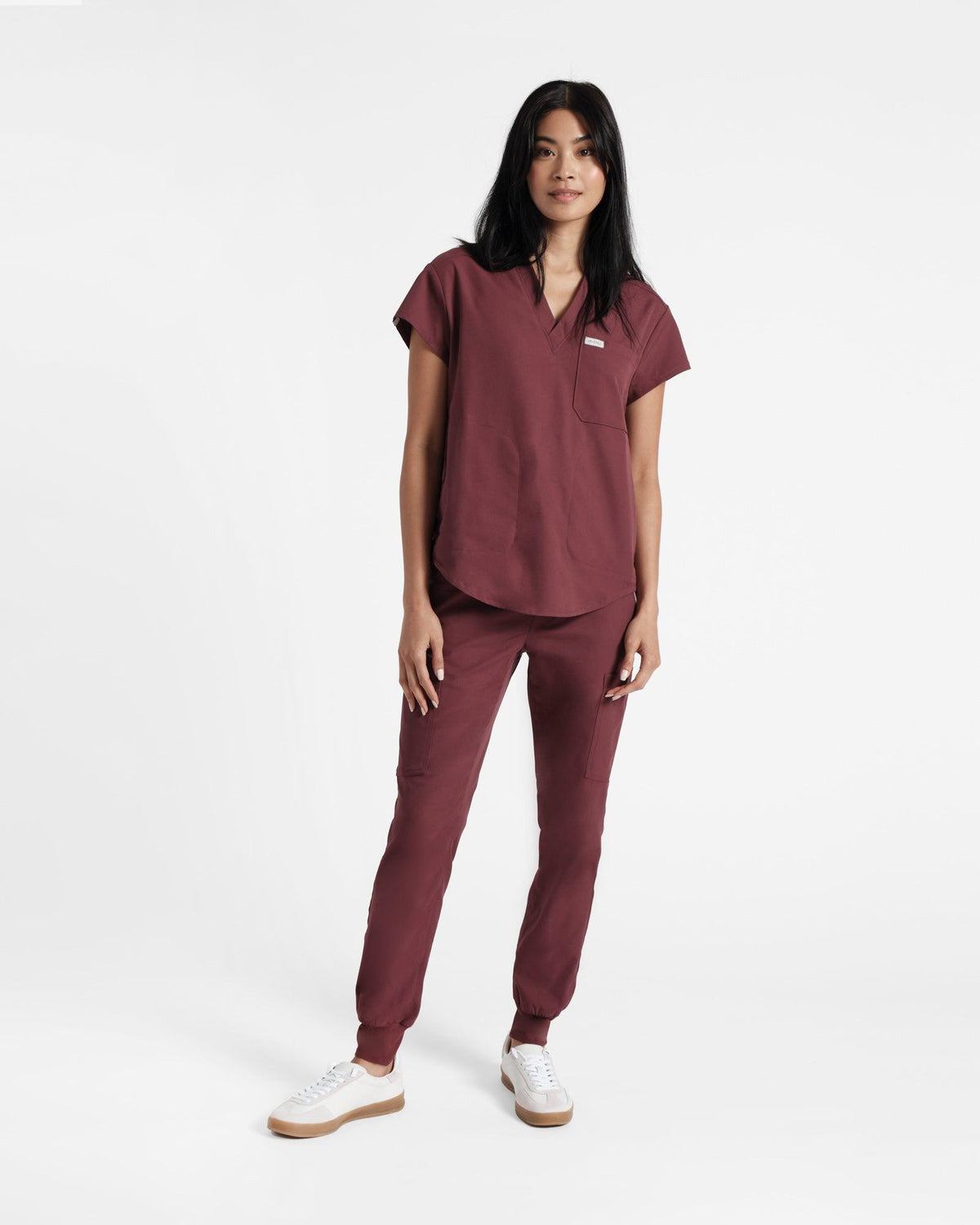 Woman wearing a deep red v-neck scrub shirt with a rounded hem, untucked over matching scrub pants.