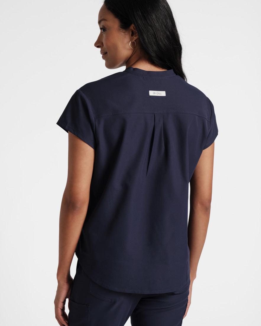 Back of woman wearing a navy scrub shirt with a back pleat and On Call fabric label on the yoke.