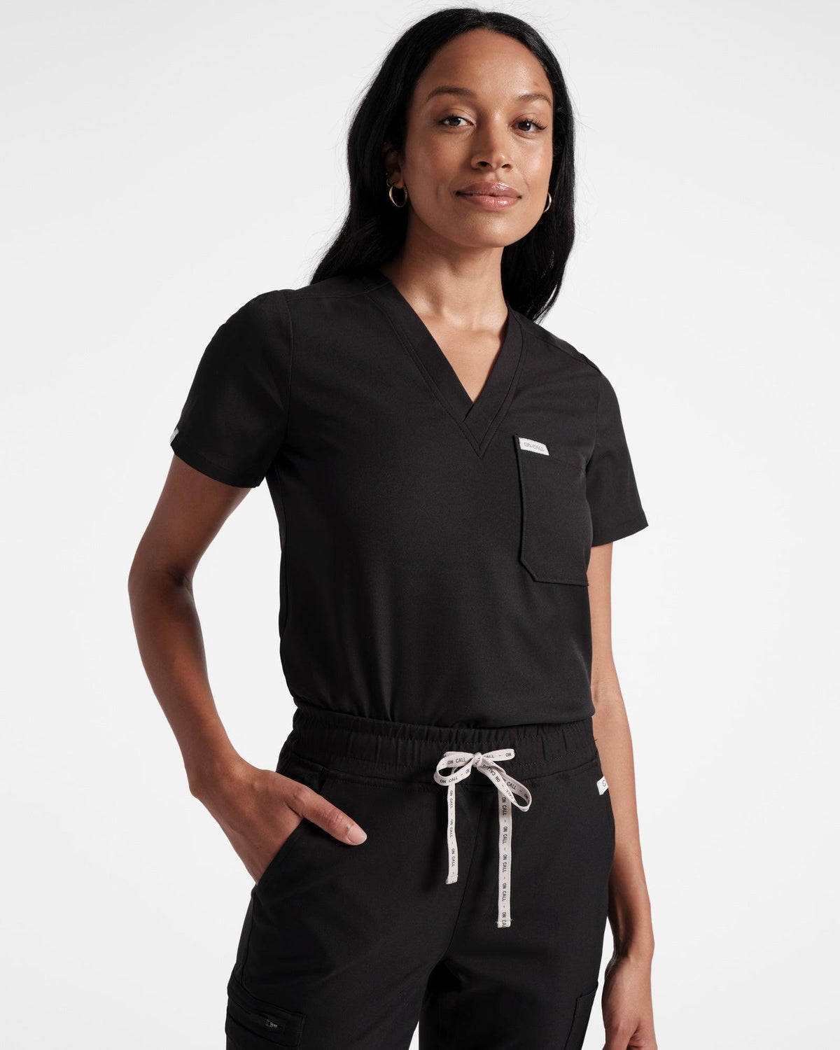 Woman wearing v-neck black scrub top with chest pocket tucked into matching scrub pants.