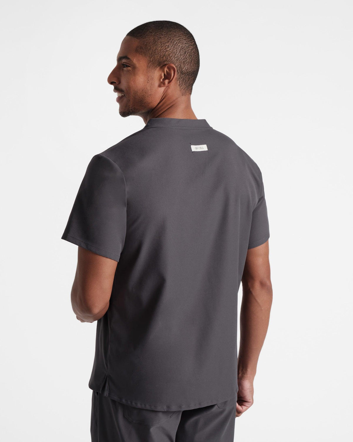 Back side view of man wearing a dark grey scrub shirt with an On Call fabric label sewn under the neck seam.