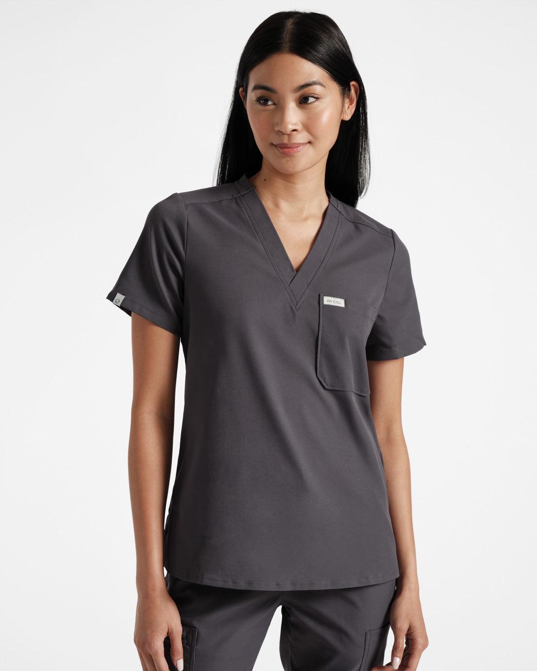 Woman in dark grey scrub shirt with a v-neck, chest pocket, and logo label wrapped around the sleeve hem.