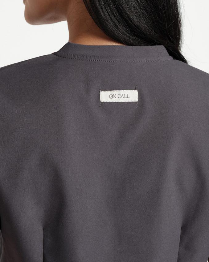 Close up back side view of a woman from the neck to mid torso in a dark grey scrub shirt with dart detailing, focusing on an On Call fabric logo label sewn into the back.