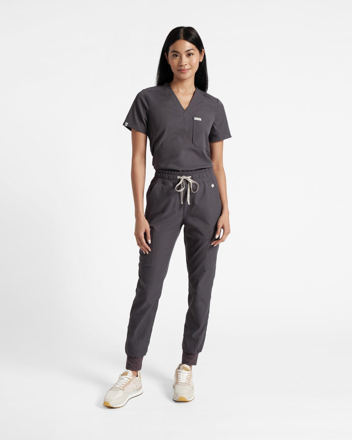 Woman in dark grey scrub shirt with a v-neck, chest pocket, and logo label wrapped around the sleeve hem, tucked into matching jogger cargo scrub pants.