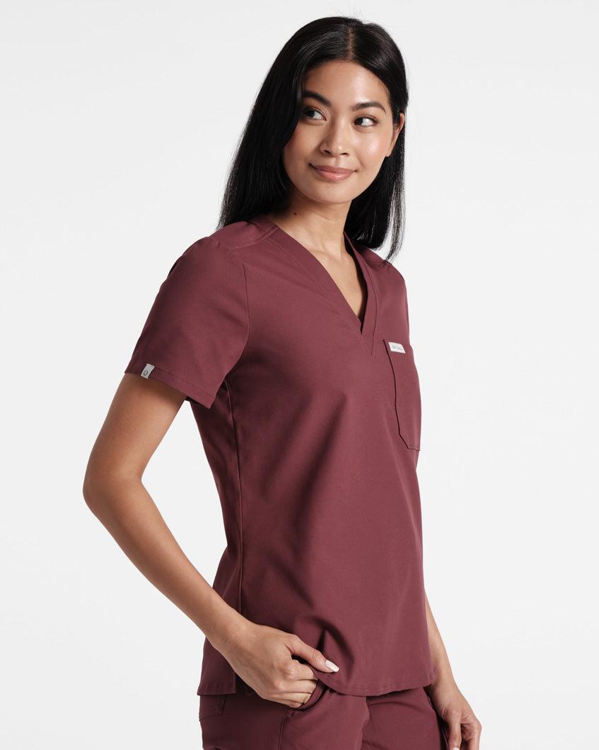 Woman in deep red scrub shirt with a v-neck, chest pocket, and small logo woven label wrapped around the sleeve hem.
