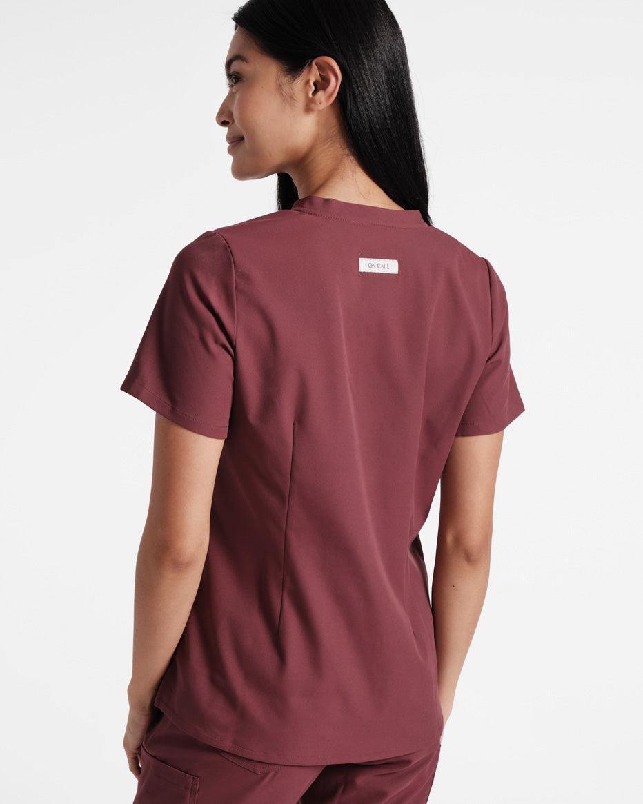 Back side view of woman in deep red scrub shirt with dart detailing and logo fabric label sewn under the neck seam.