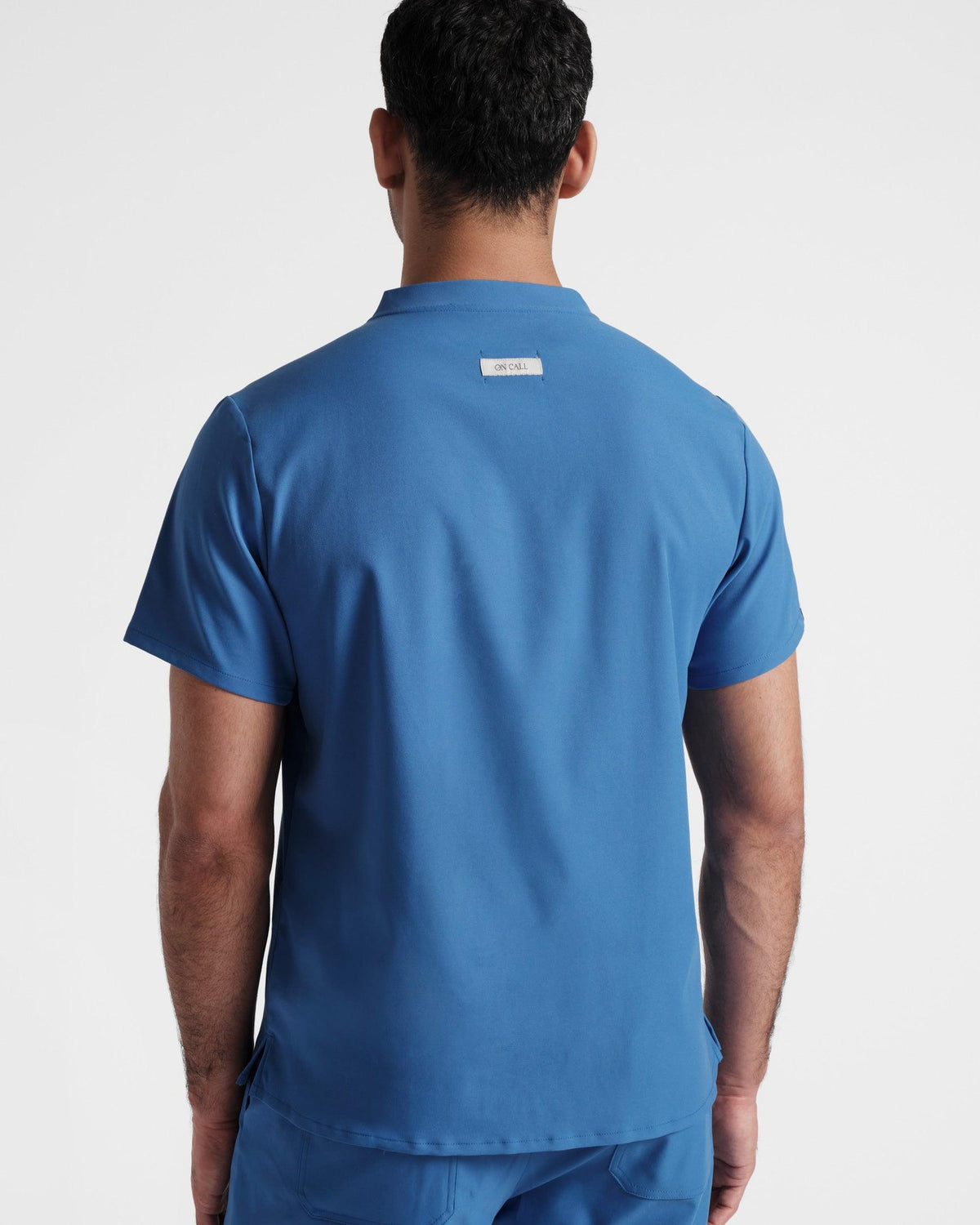 Back side view of a man wearing a bright blue scrub shirt with an On Call logo fabric label sewn under the neck seam.