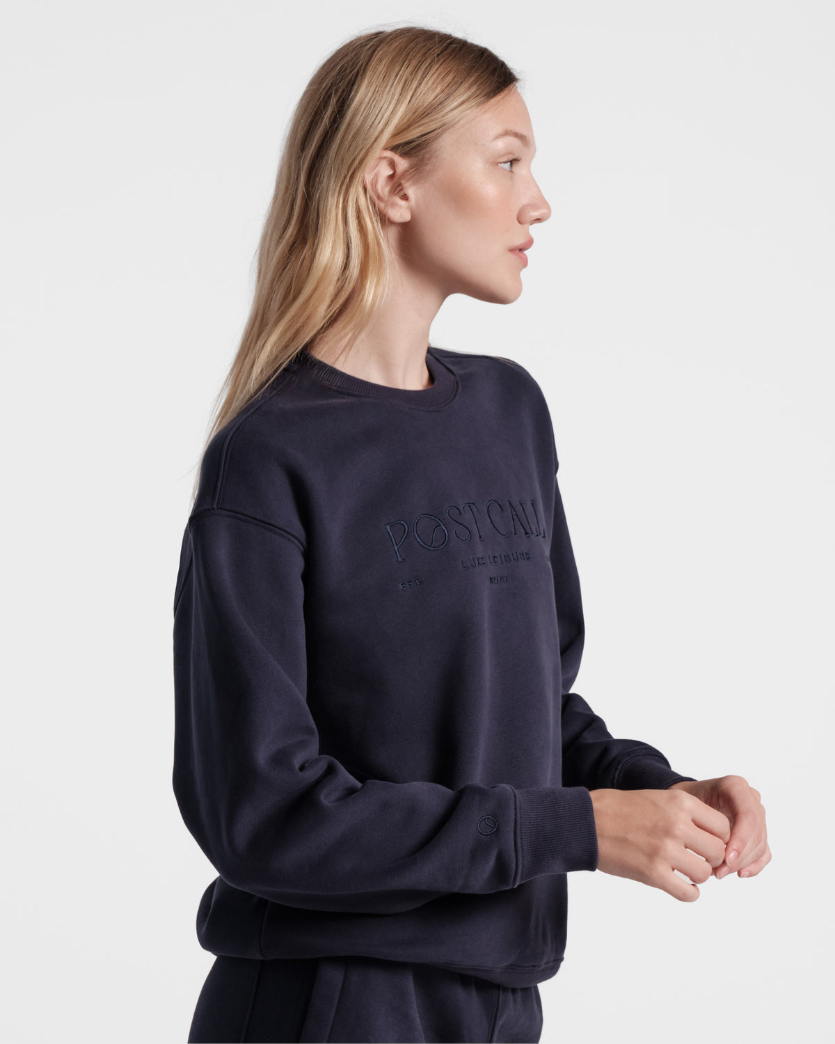 Side view of woman wearing unisex navy crewneck sweatshirt with tonal Post Call logo embroidery across the chest and small Post Call emblem logo above the wrist cuff.