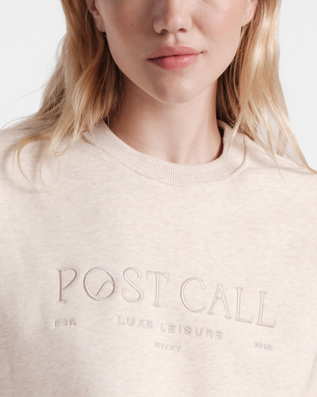 Close up of woman wearing wearing a heather beige unisex crewneck sweatshirt with Post Call tonal logo embroidery across the chest.
