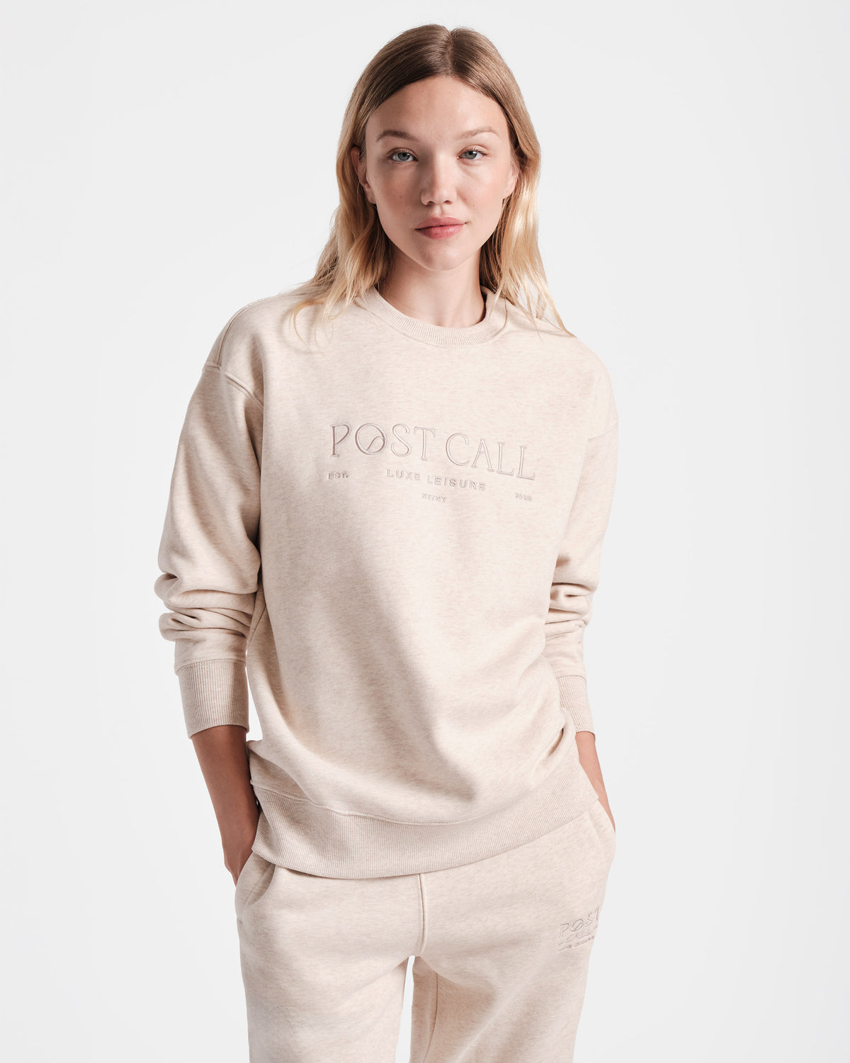 A woman wearing a unisex heather beige crewneck sweatshirt with the text 'POST CALL' on the front.