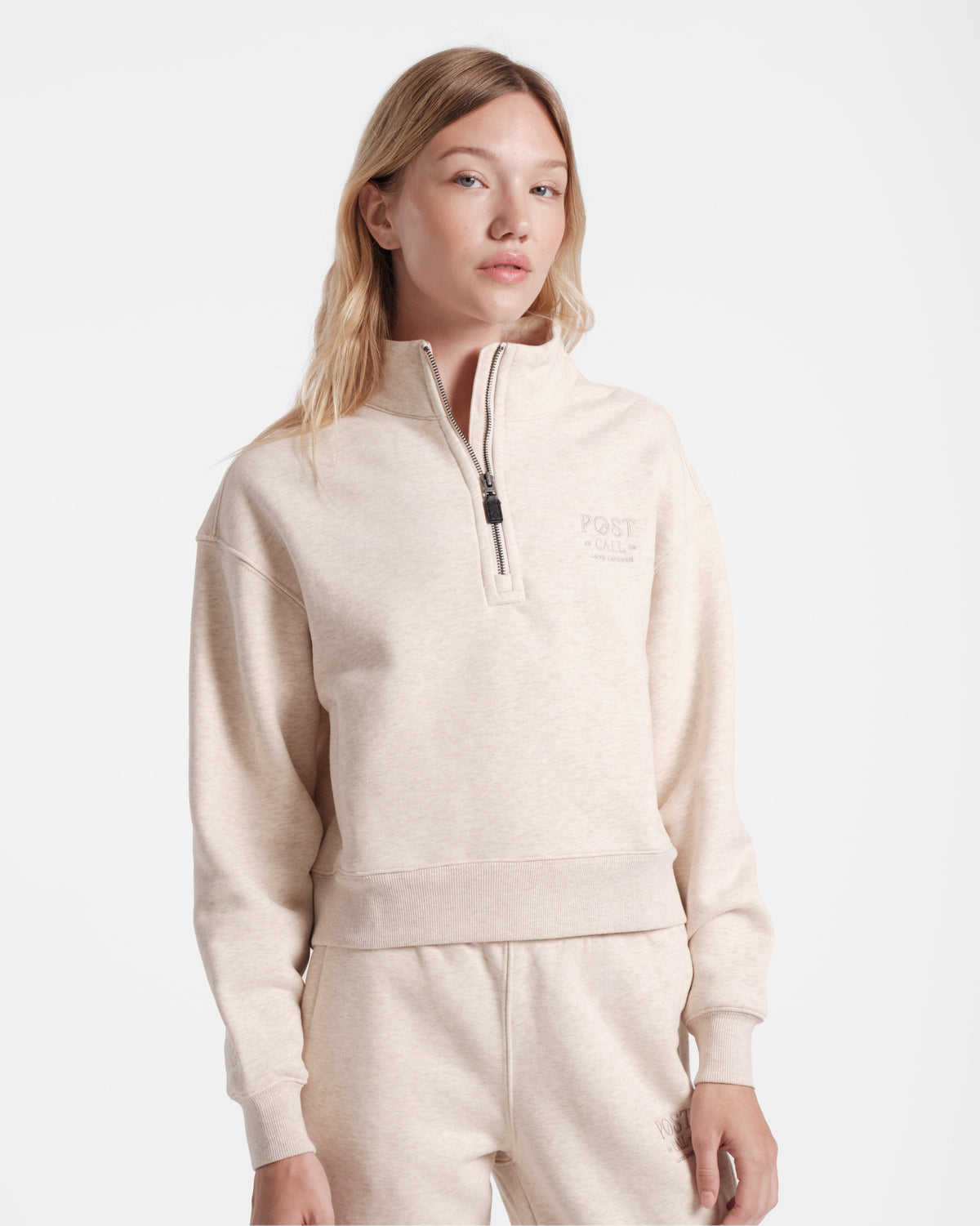 Woman wearing heather beige half zip sweatshirt with a leather zip pull and tonal Post Call logo embroidery on the left chest.