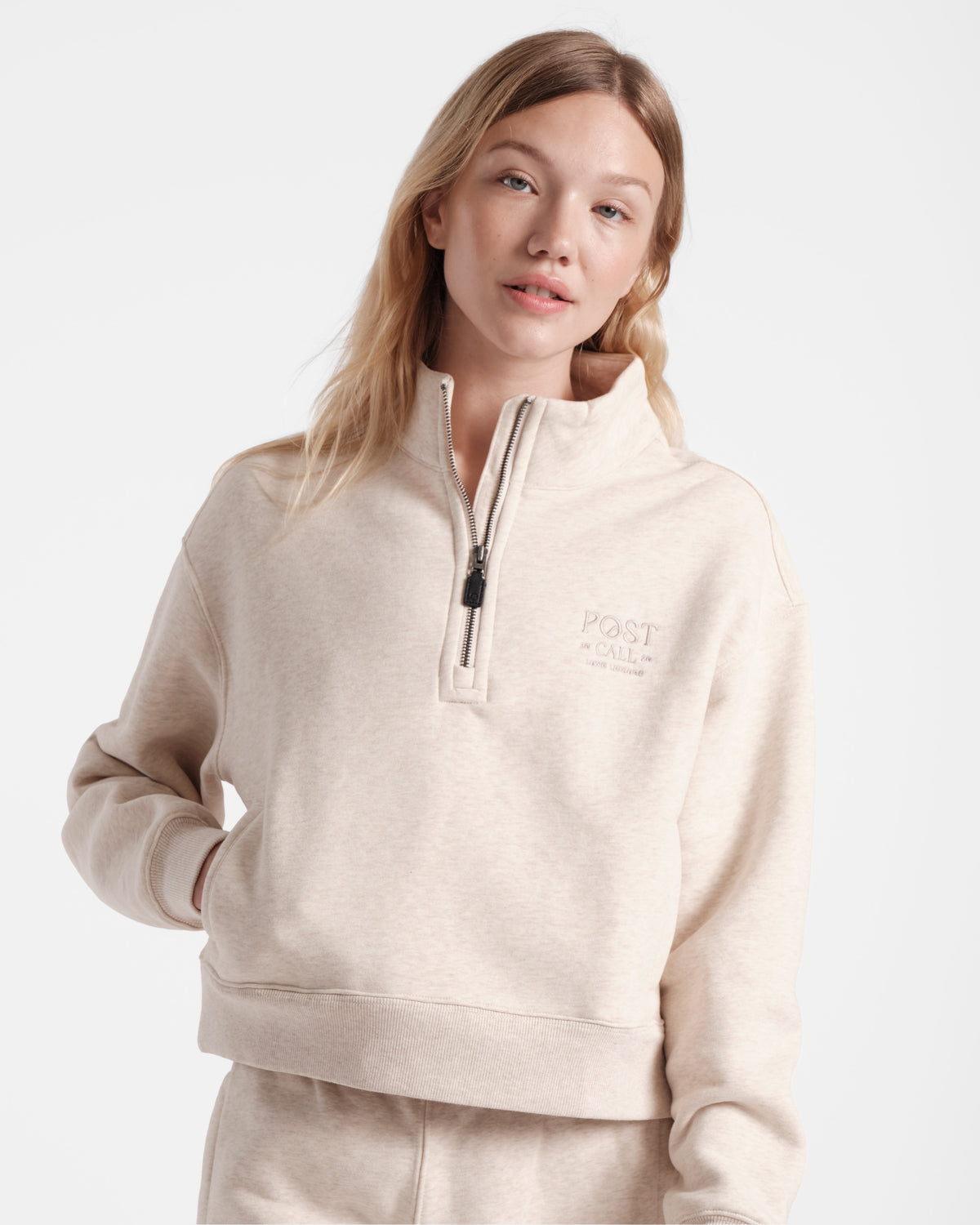 Woman wearing heather beige half zip sweatshirt with a leather zip pull and tonal Post Call logo embroidery on the left chest with her hand in one of the side pockets.