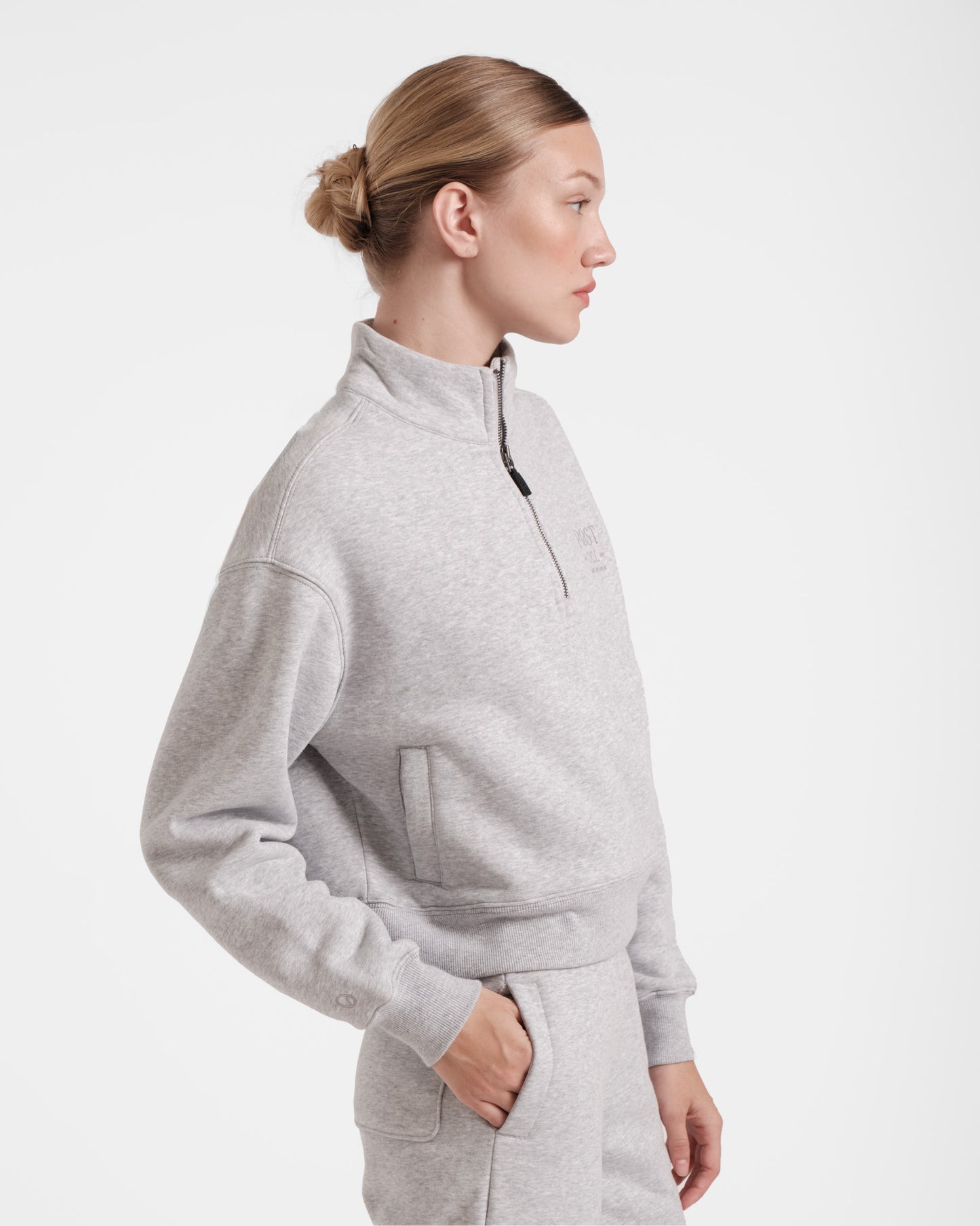 Angled side view of woman wearing heather grey half zip sweatshirt with a leather zip pull and waist pockets.