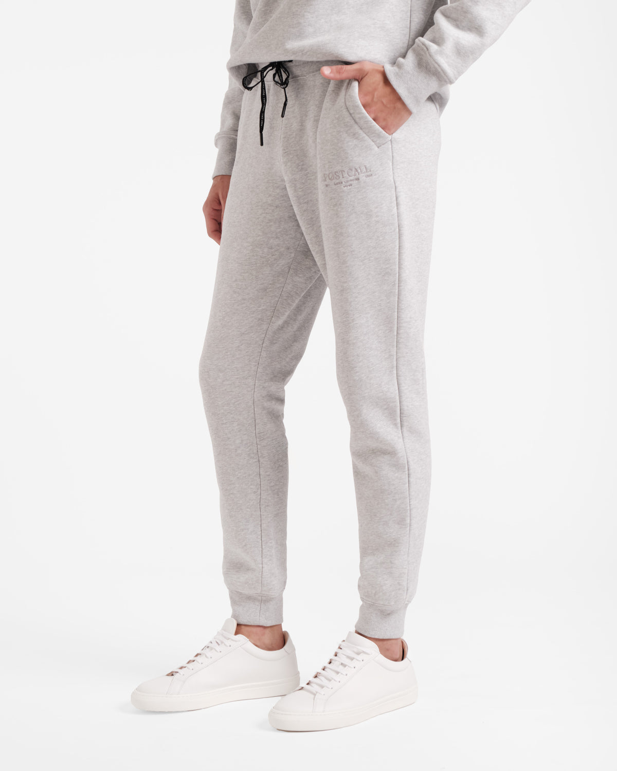 Waist down angled view of man wearing heather grey jogger sweatpants with stretch ankle cuffs, black logo branded waist tie, and tonal Post Call logo embroidery on the upper thigh.