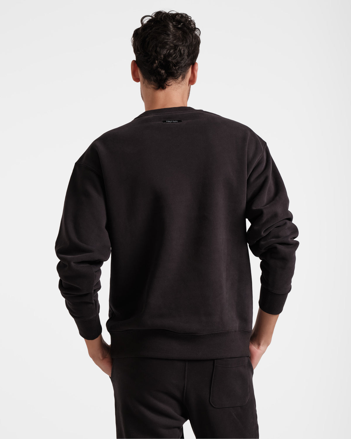 Back side view of man wearing black unisex crewneck sweatshirt with black Post Call fabric label under the neck seam.