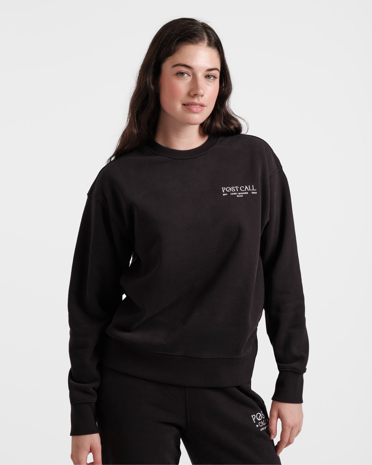 Woman wearing a unisex black crewneck sweatshirt with Post Call logo contrast embroidery on the left chest.