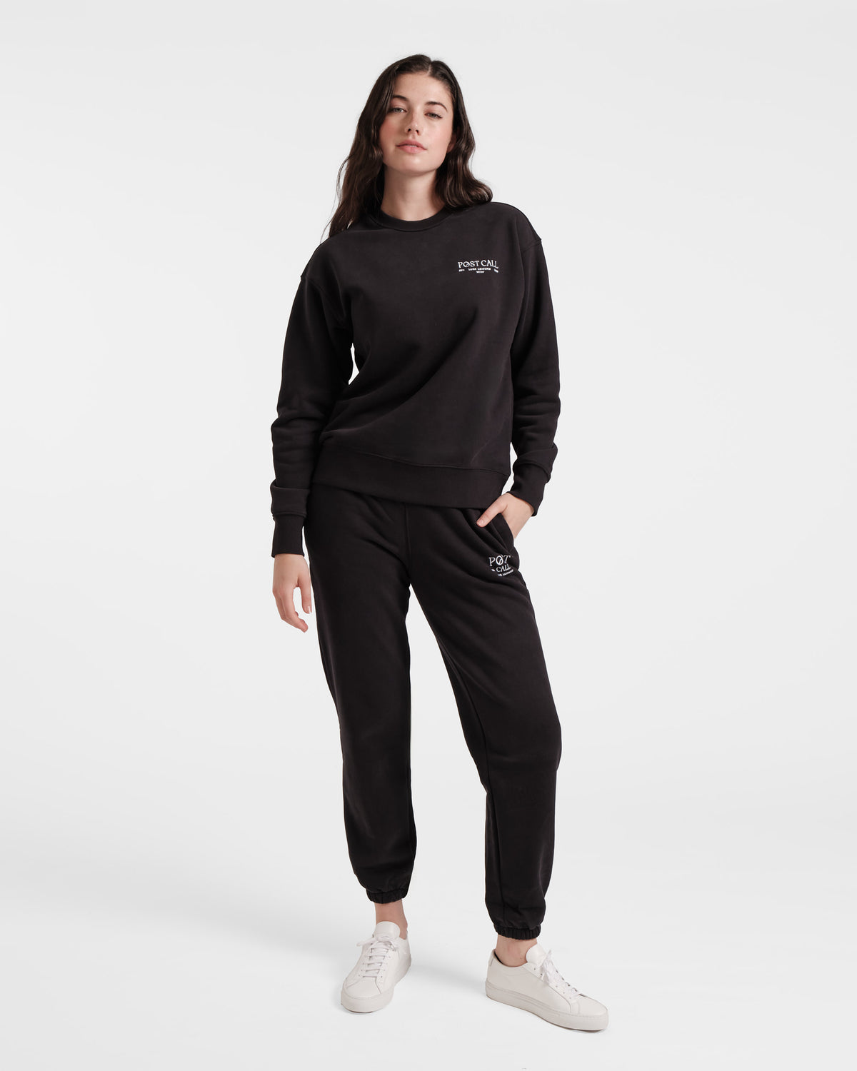 Woman wearing a matching black unisex crewneck sweatshirt and jogger sweatpants, both with white logo embroidery.