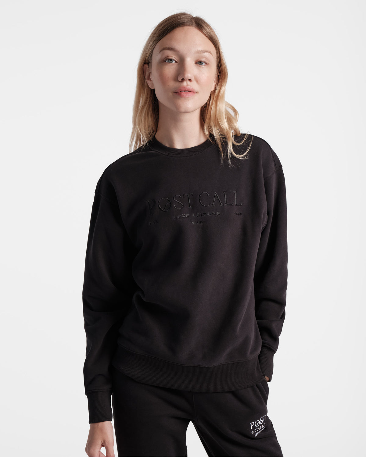 Woman wearing unisex black crewneck sweatshirt with tonal Post Call logo embroidery across the chest.