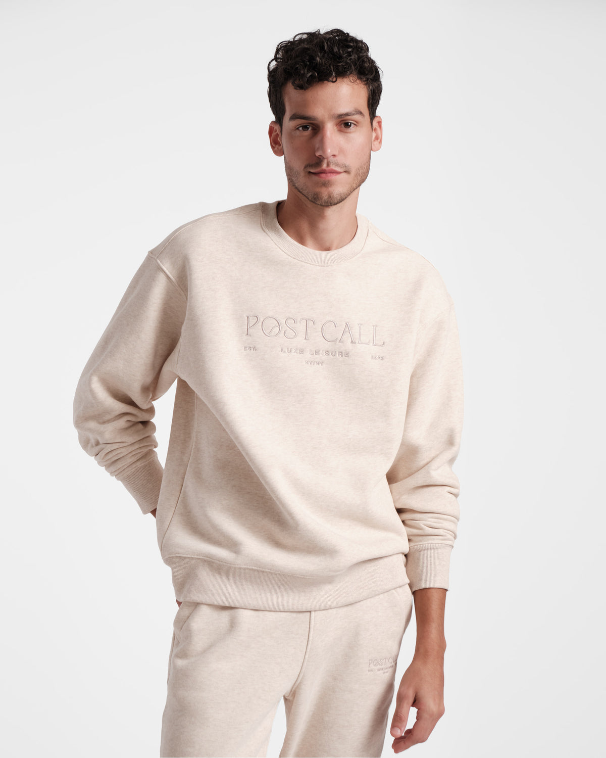 Man wearing a unisex heather beige crewneck sweatshirt with the text 'POST CALL' on the front in tonal embroidery.