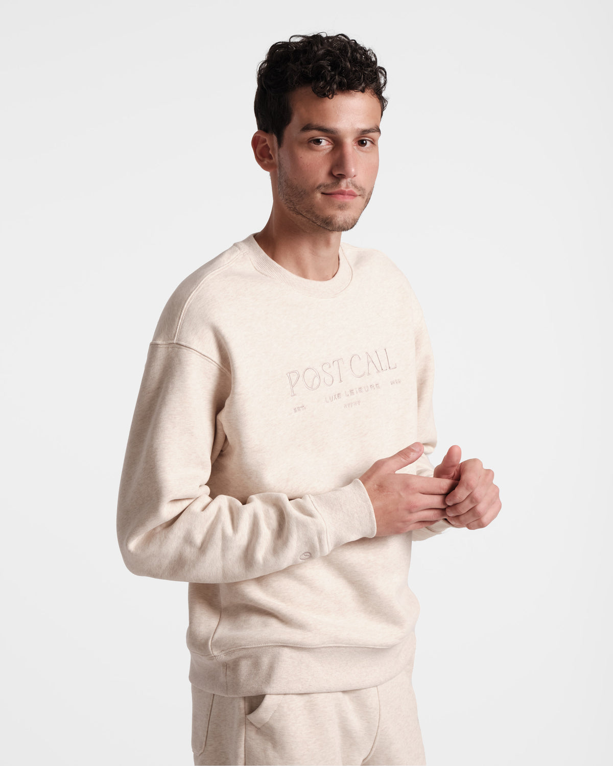 Man wearing a unisex heather beige crewneck sweatshirt with the text 'POST CALL' on the front in tonal embroidery.