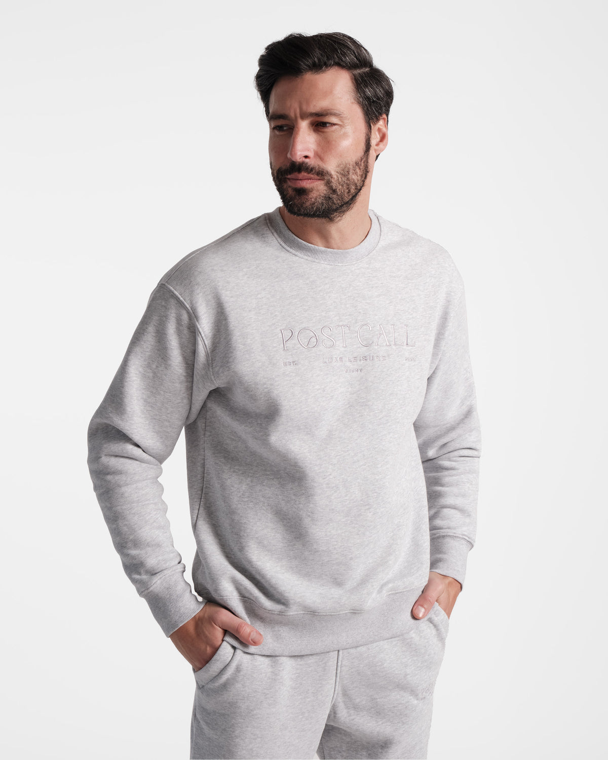 A man wearing a unisex heather grey crewneck sweatshirt with tonal Post Call logo embroidery and ribbed sleeve cuffs, standing with both hands in matching sweatpant pockets.