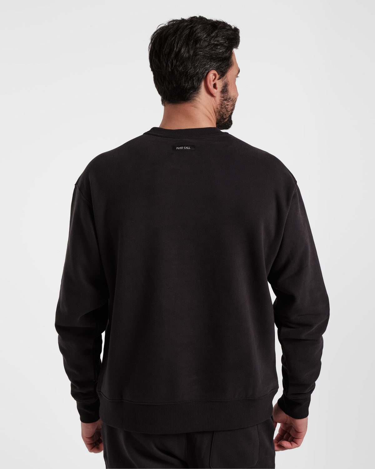 Back side view of man wearing black crewneck sweatshirt with Post Call fabric label sewn under the neck seam.