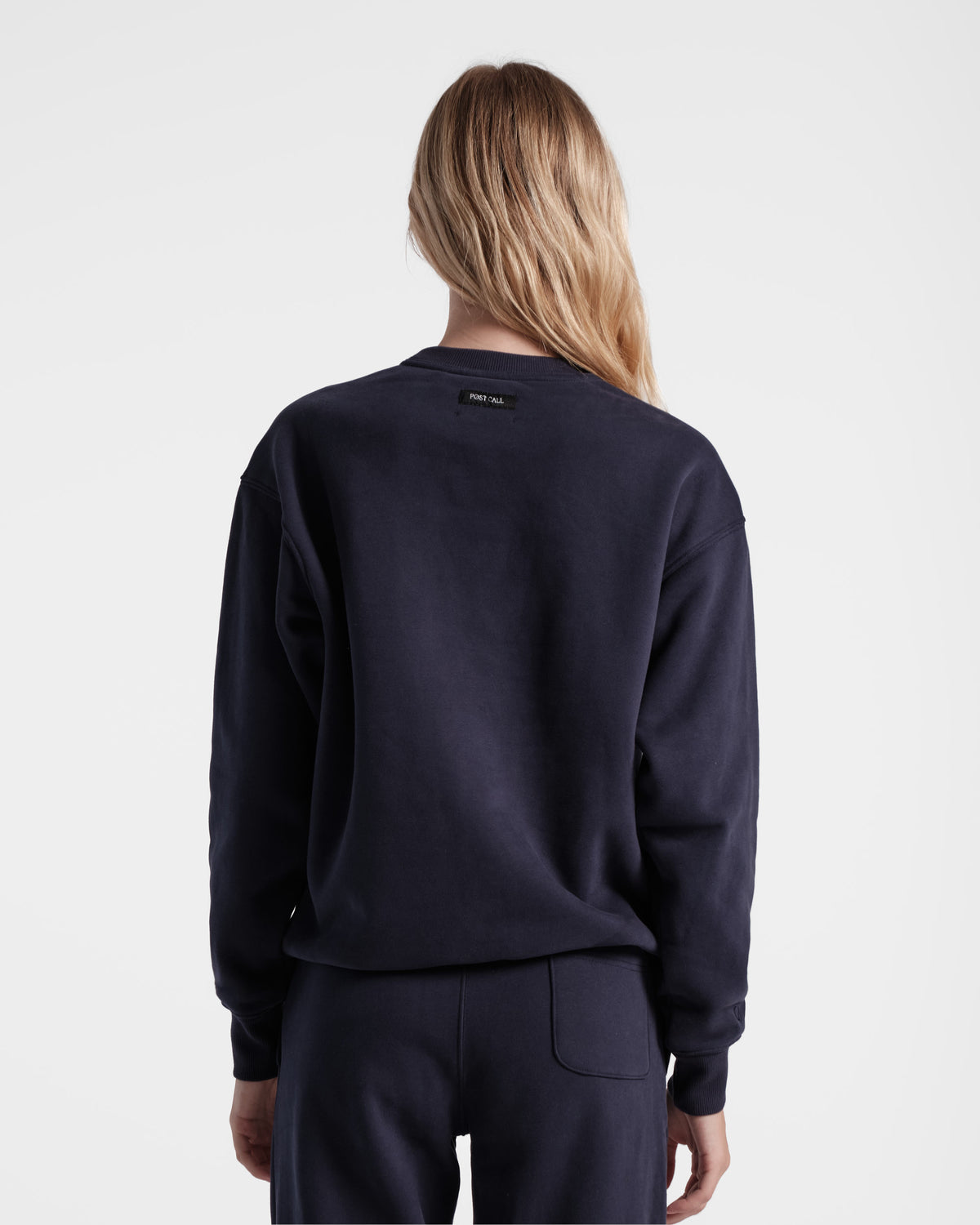 Back side view of woman wearing unisex navy crewneck sweatshirt with black Post Call fabric label sewn under neck seam.