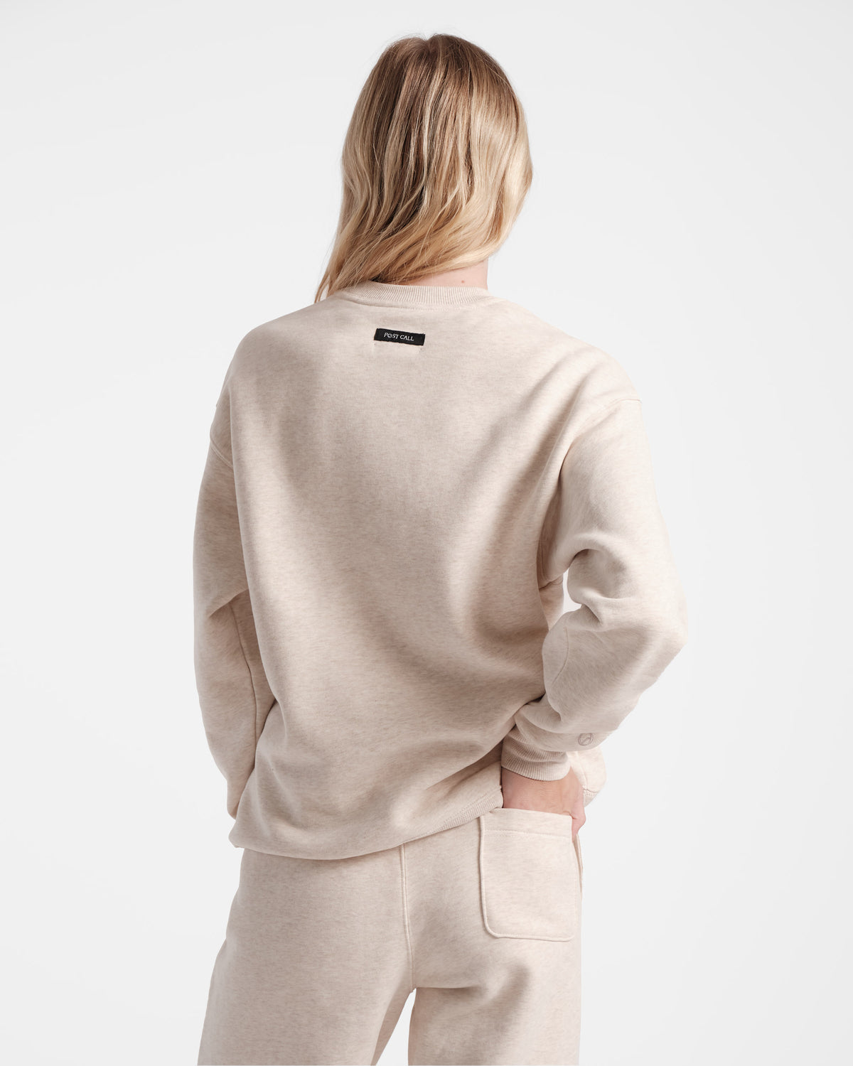 Back side view of woman wearing a heather beige crewneck sweatshirt with a black Post Call fabric label sewn under the neck seam.