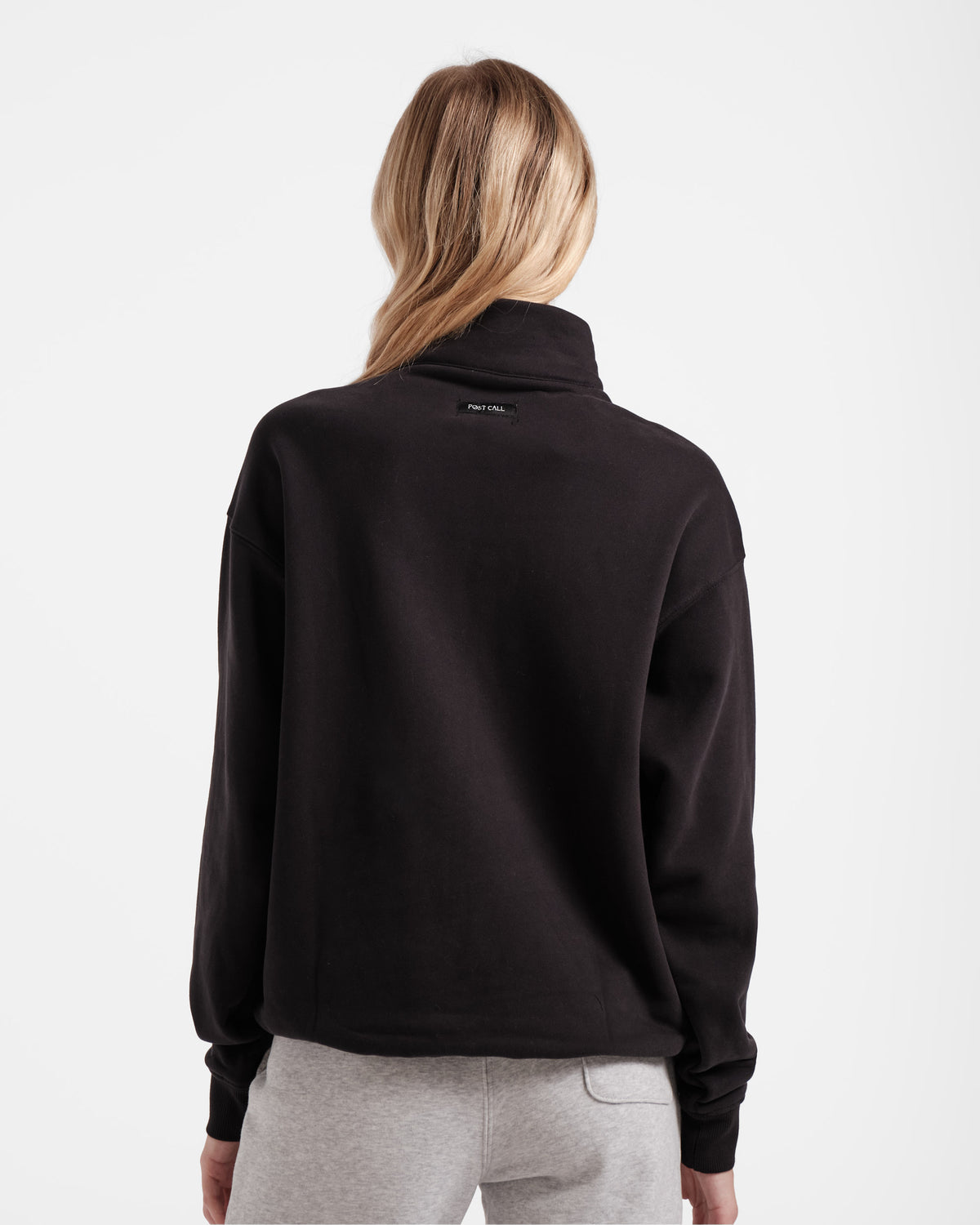Back side view of woman wearing a men's cut black half zip sweatshirt with a Post Call fabric label sewn into the back panel under the neck seam.