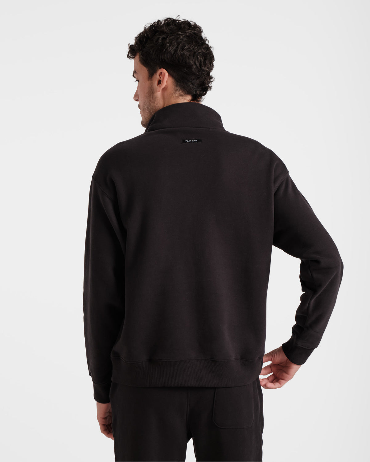 Back side view of man wearing black half zip sweatshirt with Post Call fabric label sewn under the collar on the back panel.