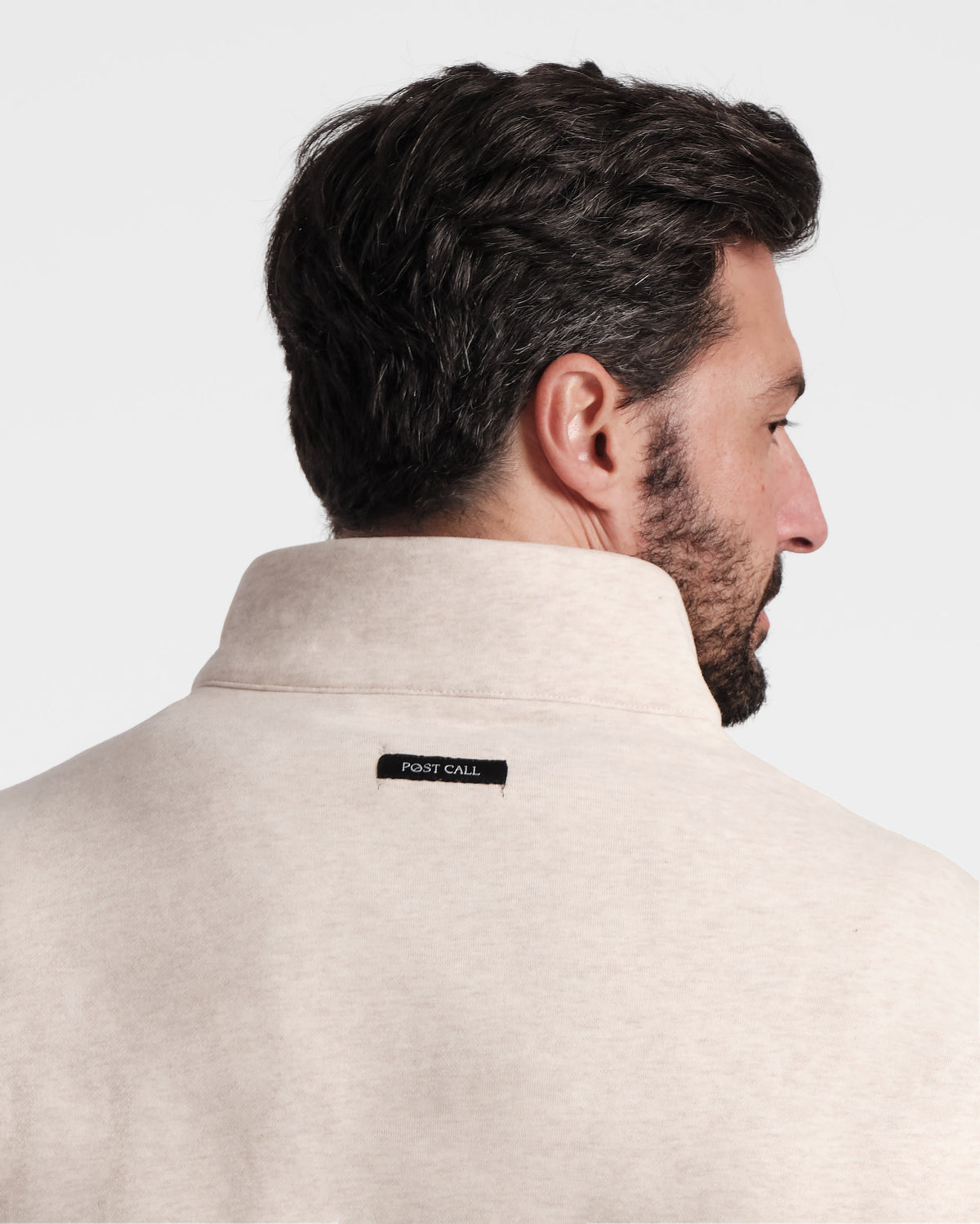 Close up back side view of man wearing heather beige half zip sweatshirt with black Post Call fabric label sewn under neck seam on the back panel.