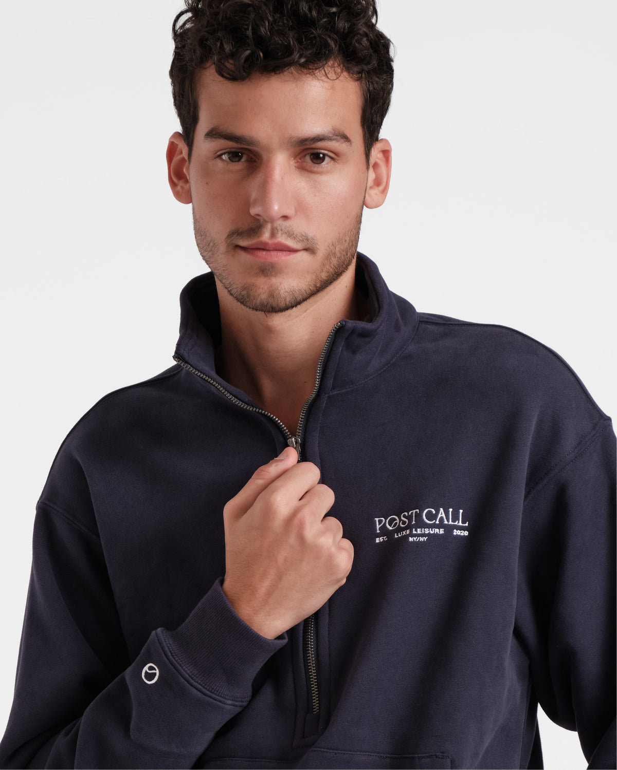 Close up view of man wearing navy half zip sweatshirt with with hand on leather zip pull, white Post Call embroidered logo on the left chest, and white Post Call emblem above wrist cuff.