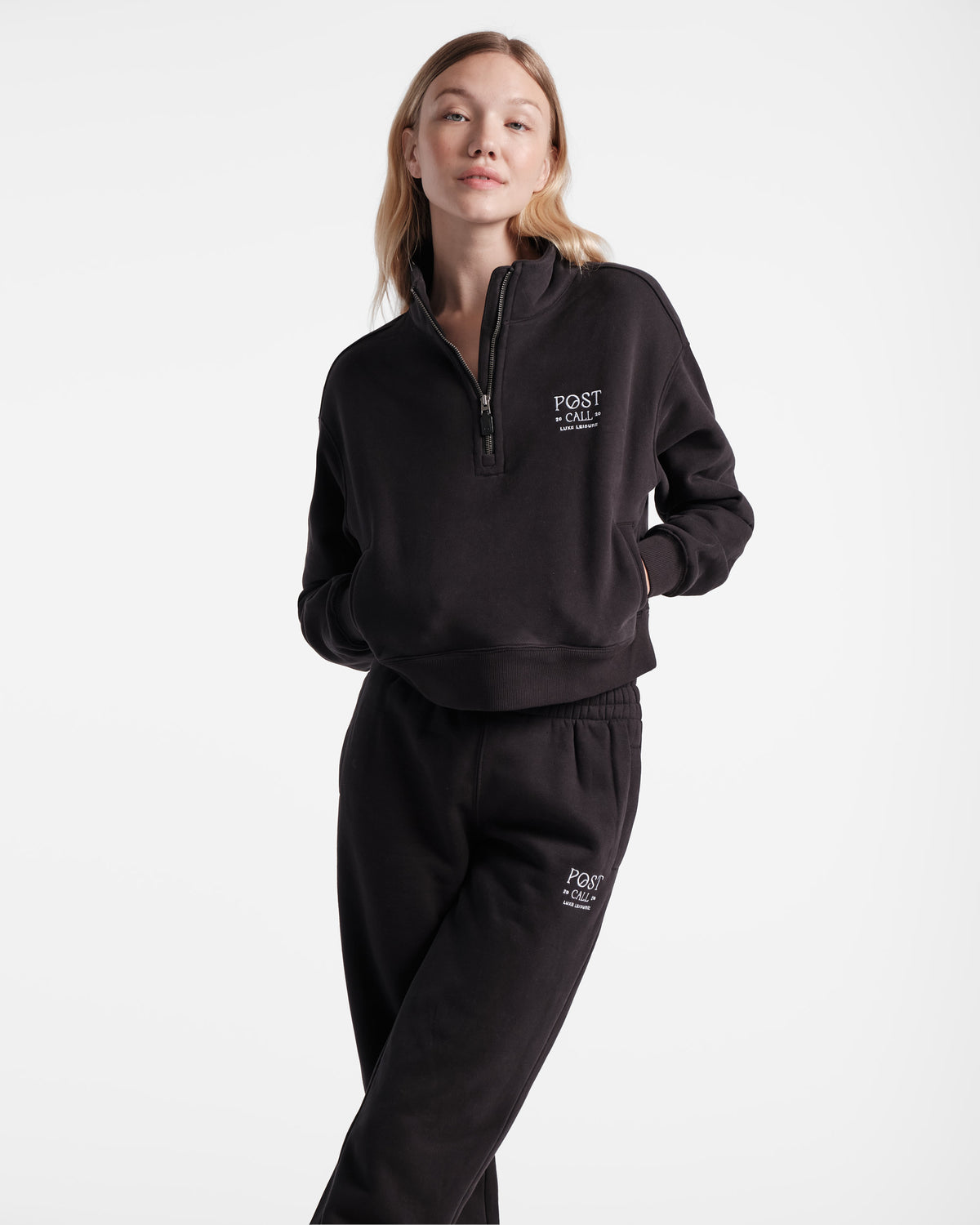 Woman wearing black half zip sweatshirt with white Post Call embroidery, hands in side pockets and legs crossed in matching sweatpants.