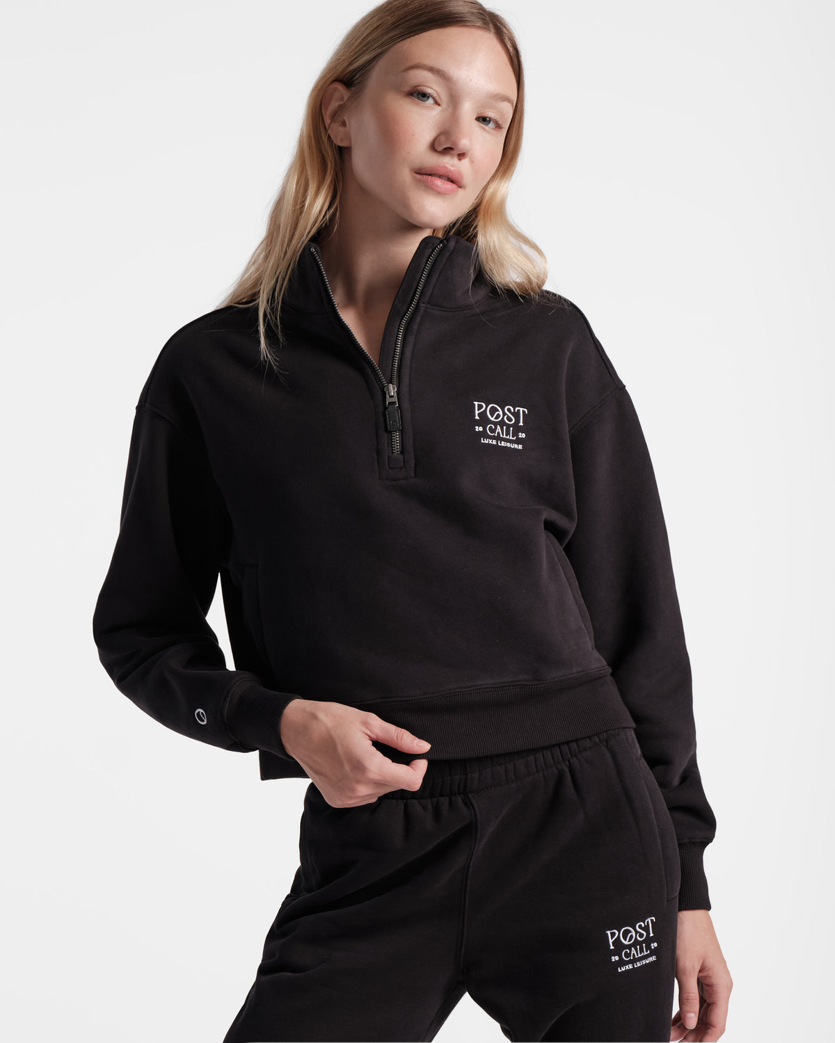Woman wearing black half zip sweatshirt with white Post Call embroidered logo on the left chest side, a small logo emblem on the right arm above the wrist cuff, and matching sweatpants.