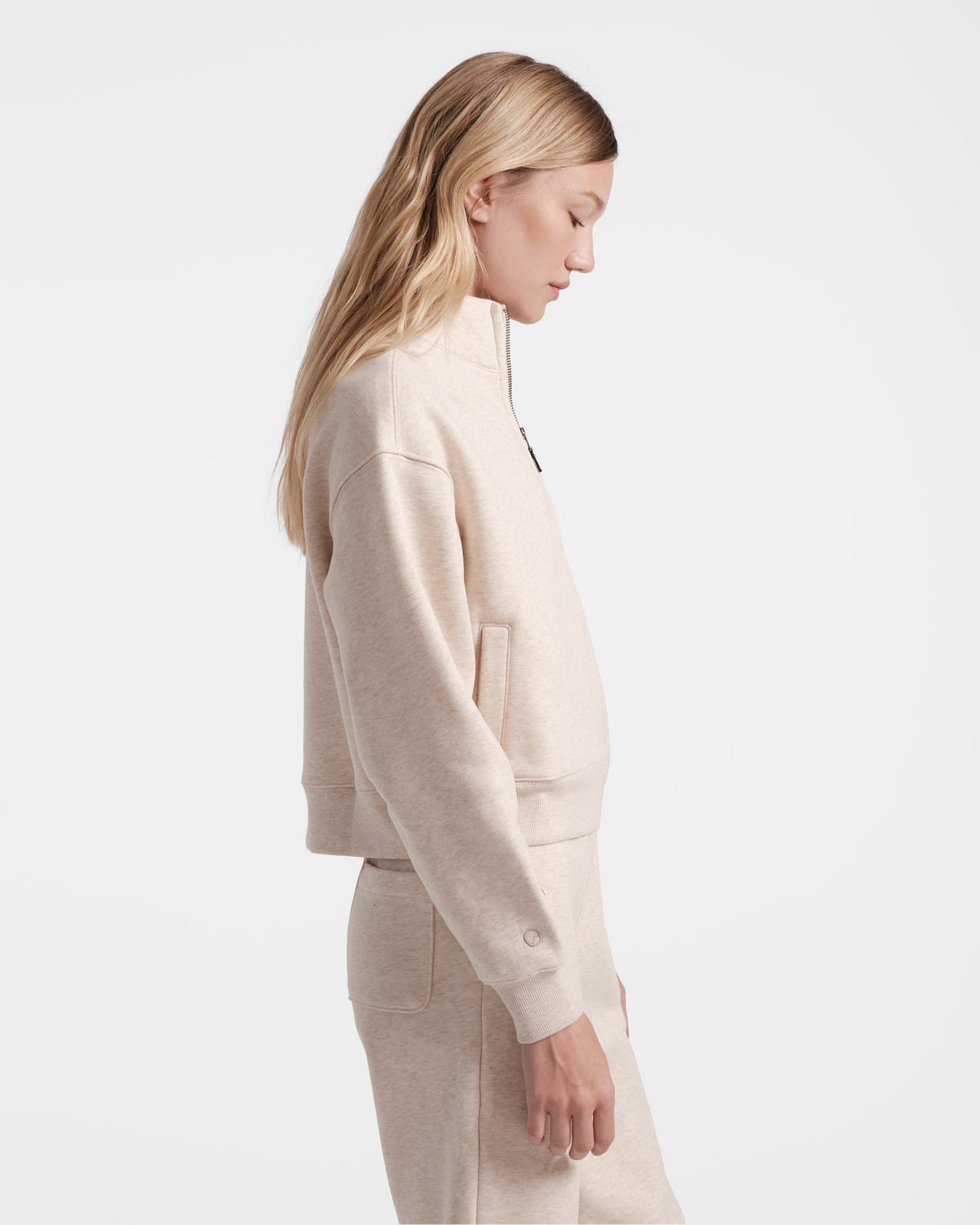 Side view of woman wearing a heather beige half zip with side pockets and tonal logo embroidery above the wrist cuff.
