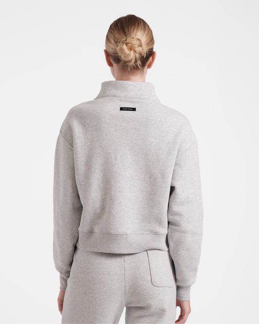 Back side view of woman wearing heather grey half zip sweatshirt with a black Post Call fabric label sewn under the neck seam on the back panel.