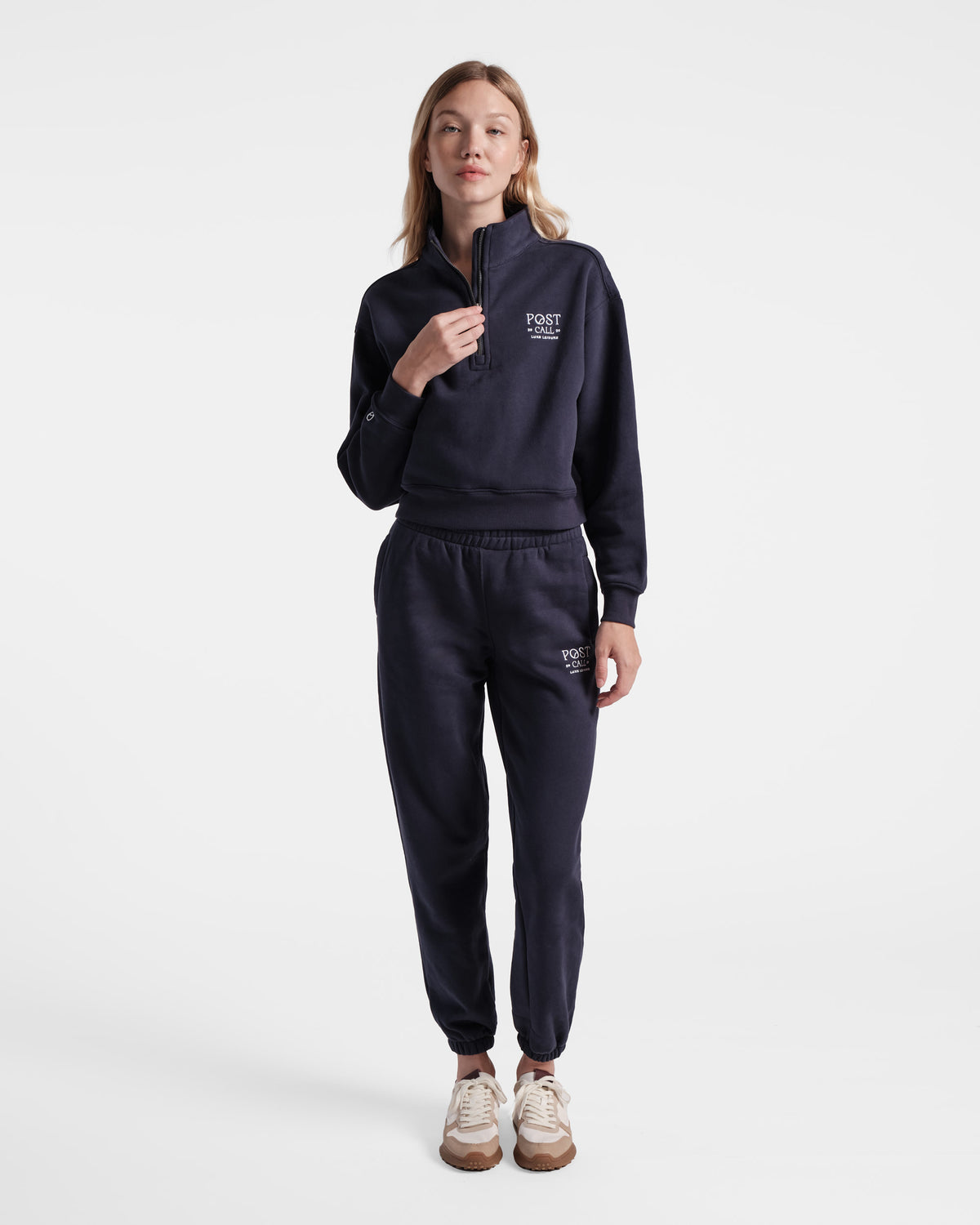 Woman wearing navy half zip sweatshirt with white Post Call logo embroidered on the left chest and hand on leather zip pull.