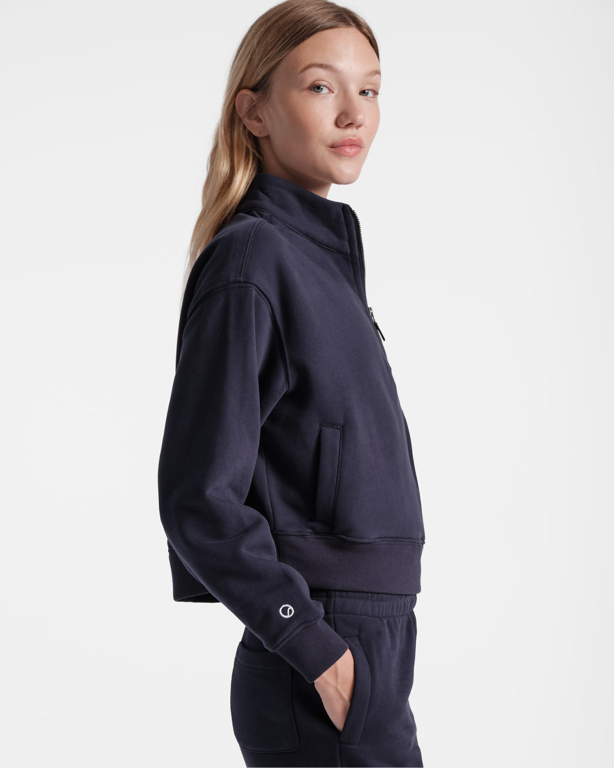 Side view of woman wearing navy half zip sweatshirt with small white embroidered logo above wrist cuff and hand in matching sweatpant pocket.