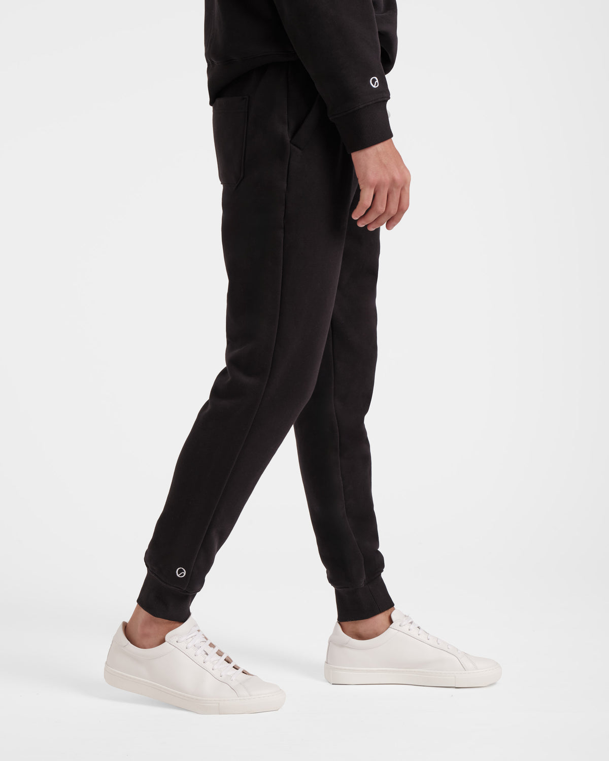 Side view of man wearing black jogger sweatpants with white small logo emblem above ankle cuff.