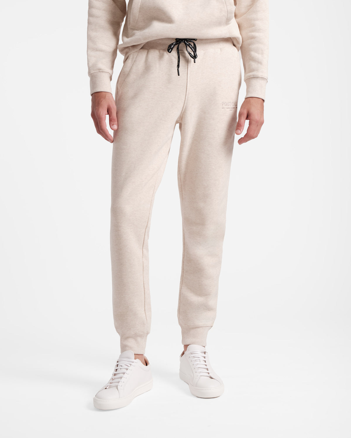 Waist down view of man wearing heather beige jogger sweatpants with stretch ankle cuffs and black contrast branded waist tie.