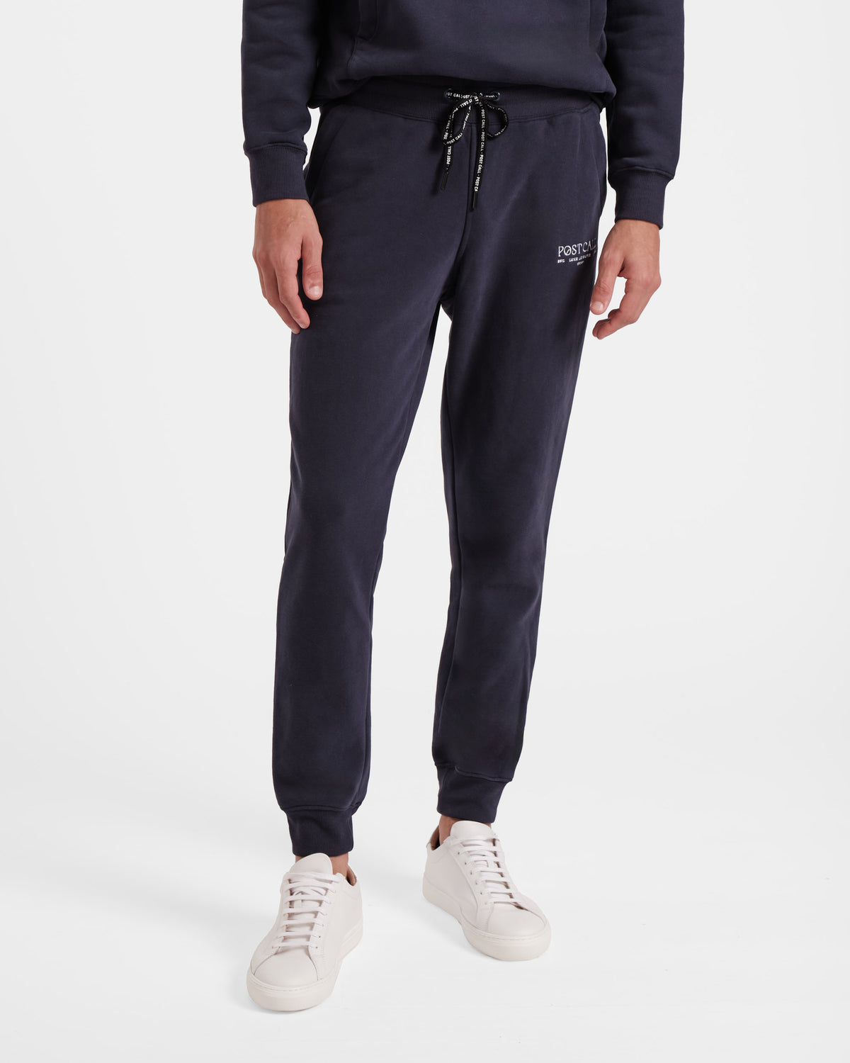 Waist down view of man wearing navy jogger sweatpants with stretch ankle cuffs, black contrast branded waist tie, and white embroidered Post Call logo on the thigh.