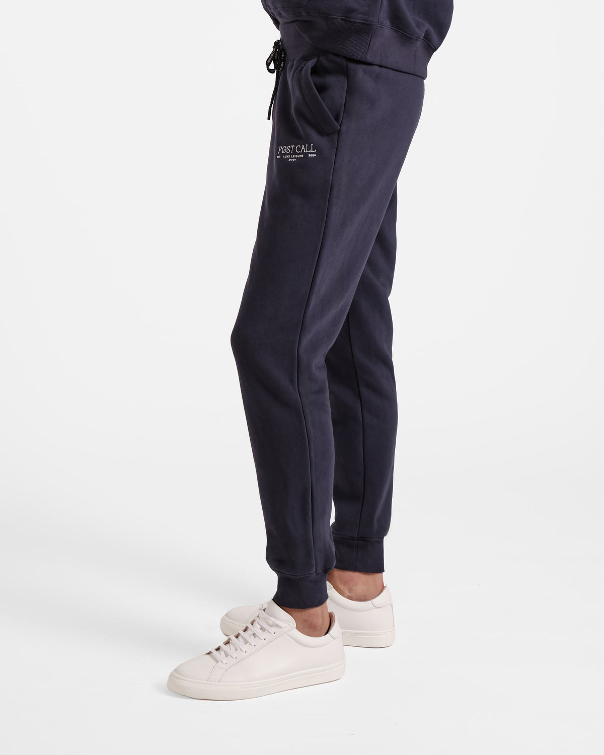 Side view of man wearing navy jogger sweatpants with stretch ankle cuffs and white embroidered Post Call logo above side pocket.