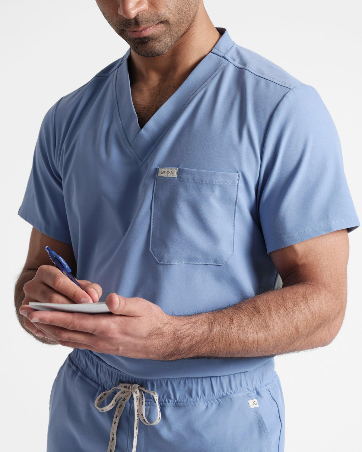 Close up view of man from the hips up in a light blue v-neck scrub shirt tucked into matching pants while writing on a notepad in hand.