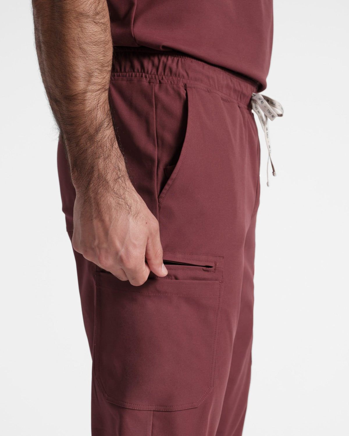 Zoomed in side view of man in deep red cargo scrub pants with hand pulling on the zipper compartment of the side leg pocket.