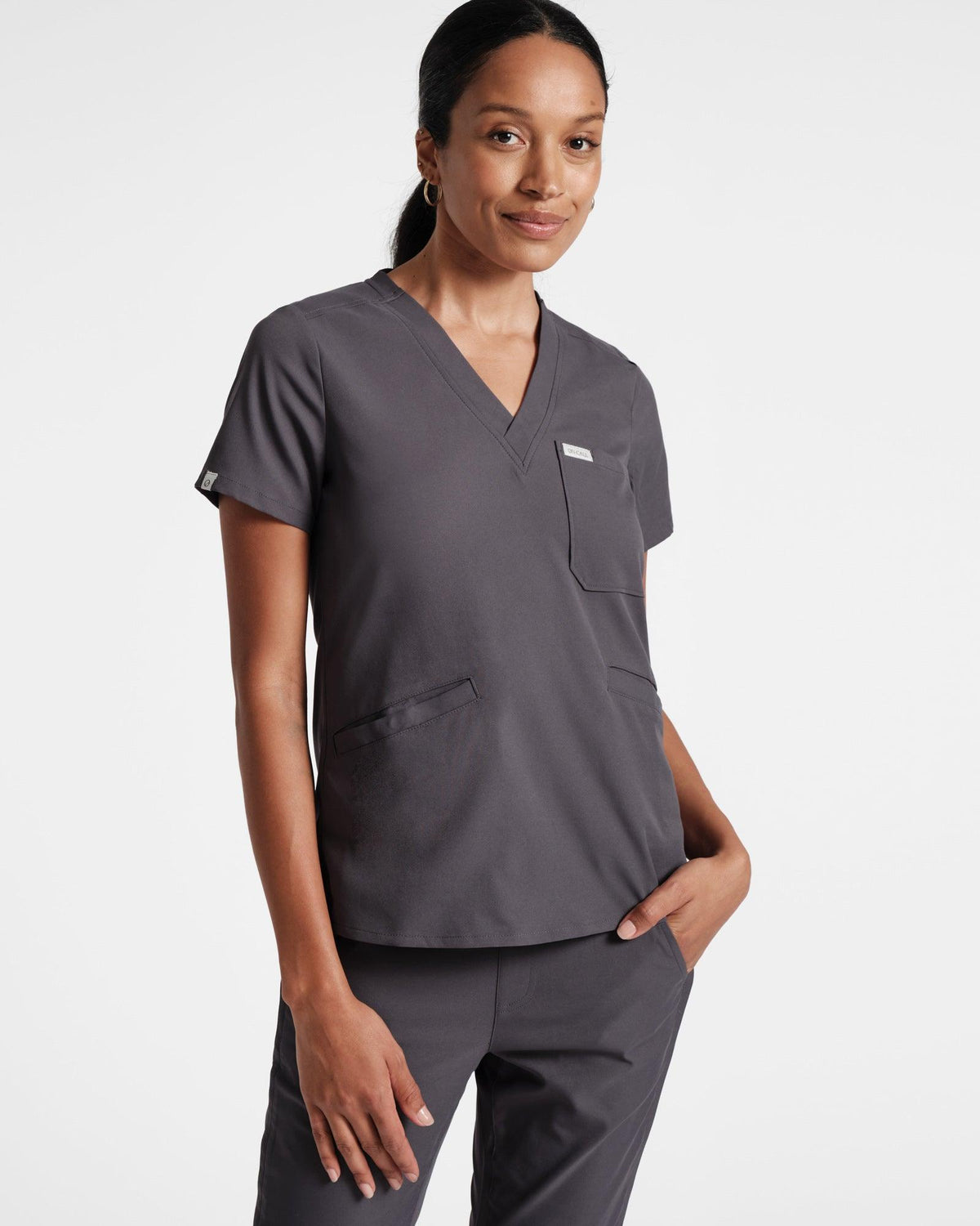 Woman wearing a dark grey scrub top with a v-neck and two front pockets.