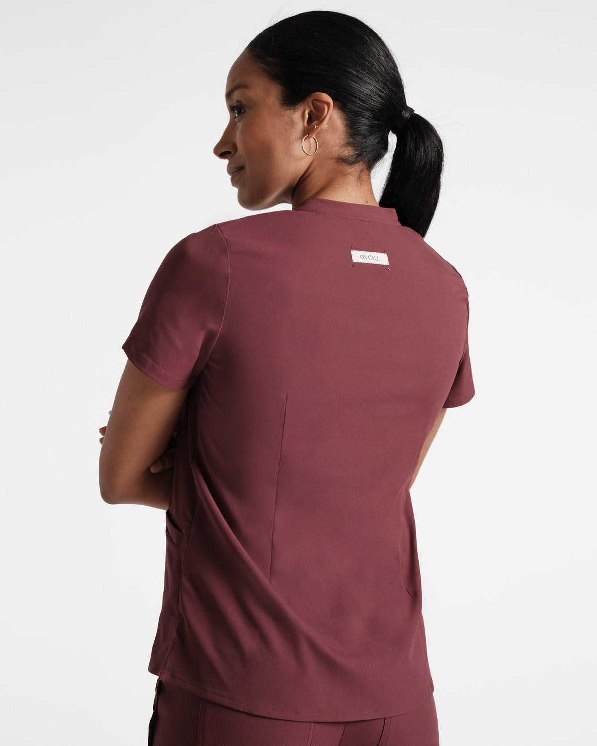 Back side view of woman in a deep red scrub shirt with dart detailing and an On Call fabric label sewn under the neck seam.