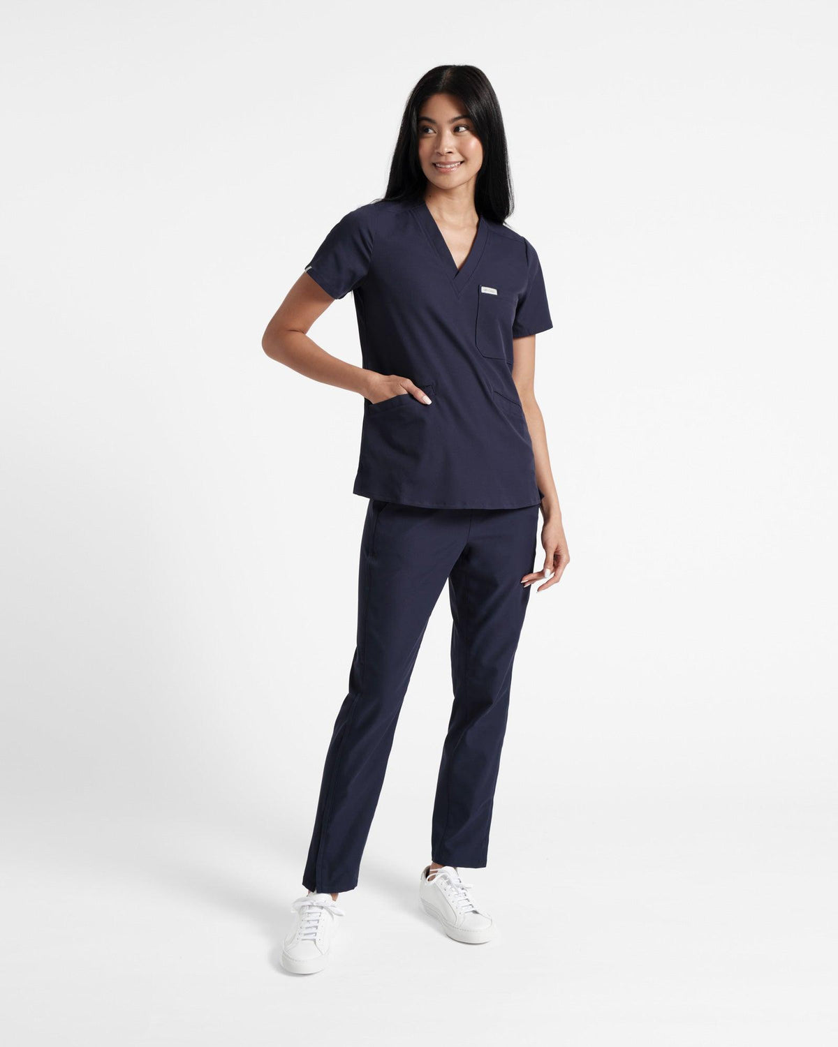 Woman wearing a navy v-neck scrub shirt with a chest pocket, two front pockets and matching pants.