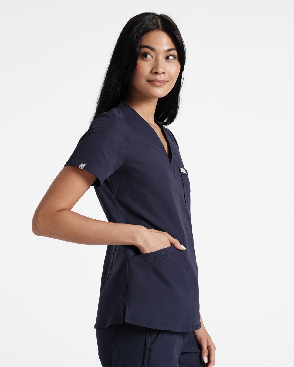 Side view of woman wearing a navy v-neck scrub shirt with a chest pocket and her hand in one of the front pockets.