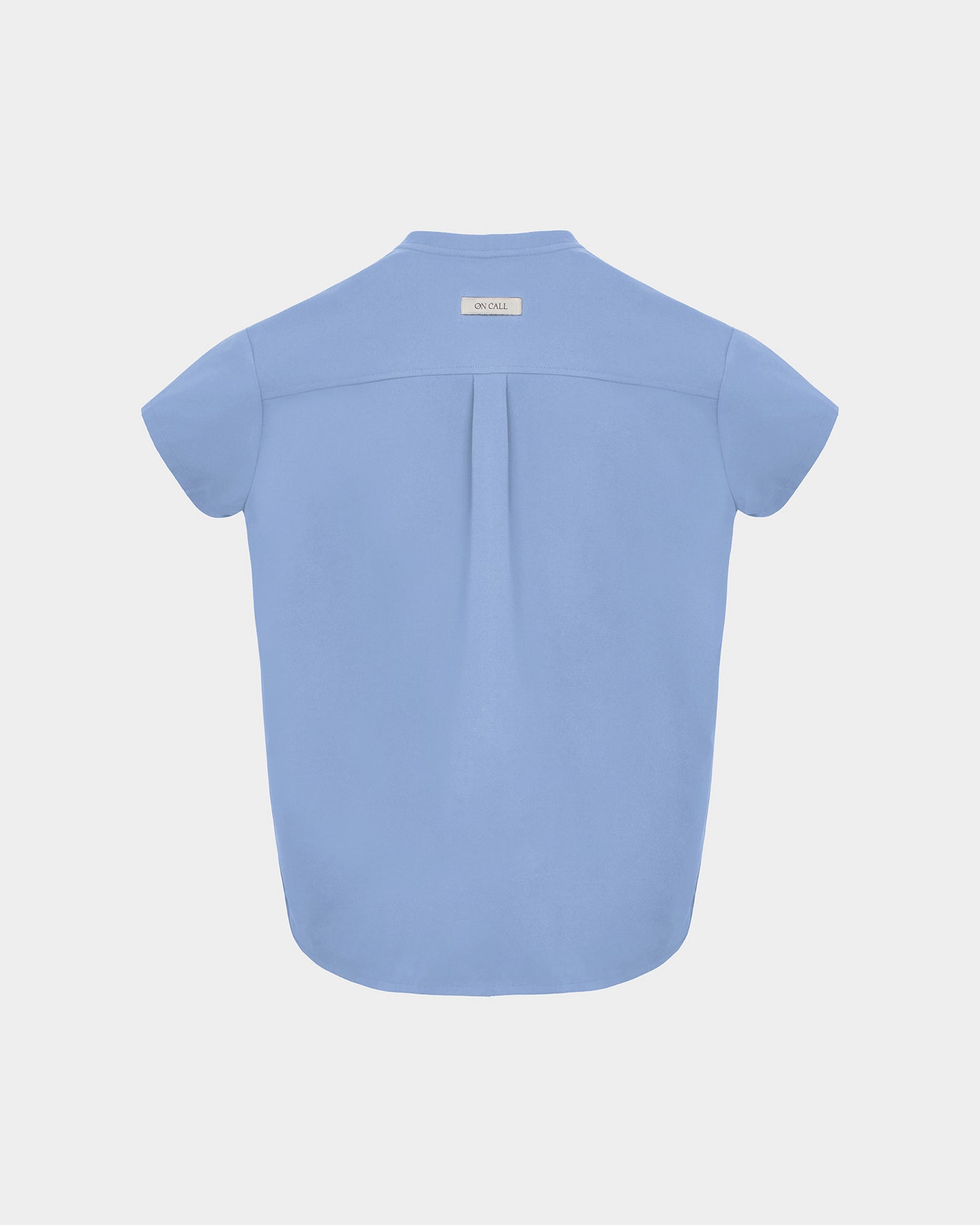 A light blue short-sleeve scrub shirt shown from the back, featuring a subtle center back pleat and a small rectangular brand label at the neckline, set against a solid light gray background.