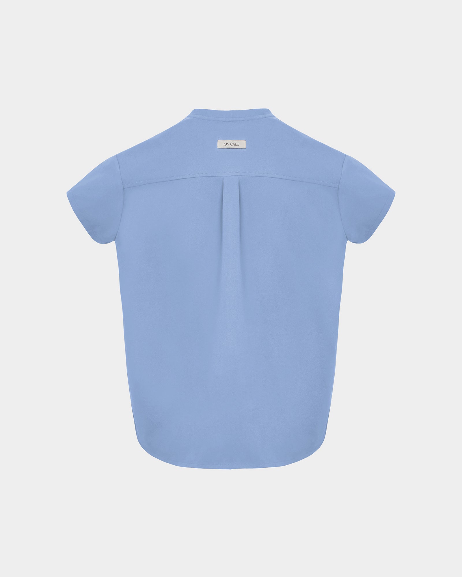 A light blue short-sleeve scrub shirt shown from the back, featuring a subtle center back pleat and a small rectangular brand label at the neckline, set against a solid light gray background.